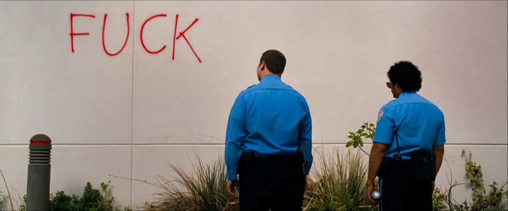 Observe And Report (2009) movie still: fullbody — A plain white exterior wall features large red spray-painted text spelling a swear word. Two…