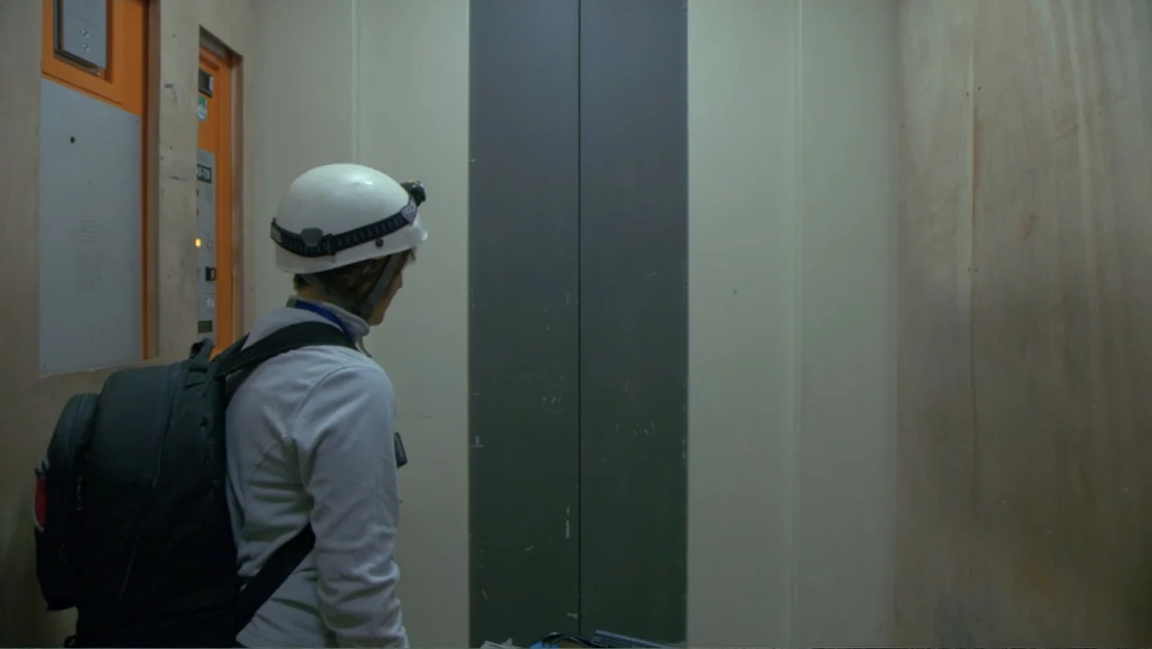 CERN (2013) movie still: medium — An interior elevator cabin with plywood-paneled walls and grey closed metal doors. A person wearing a white safety…