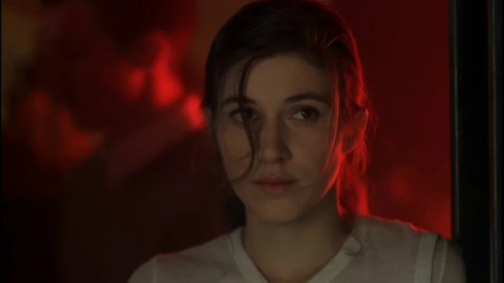 Romance (1999) movie still: closeup — A dimly lit room bathed in an intense red neon glow at night. A young woman stands looking toward the side of…