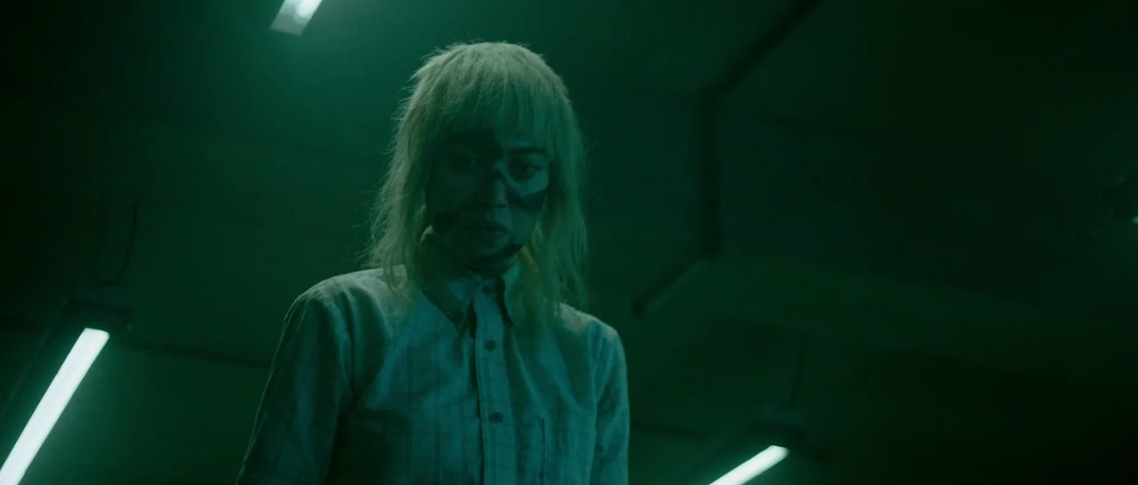 Green Room (2015) movie still: medium — A dimly lit room is filled with a distinct green-tinted atmosphere. A young woman with dark facial markings…