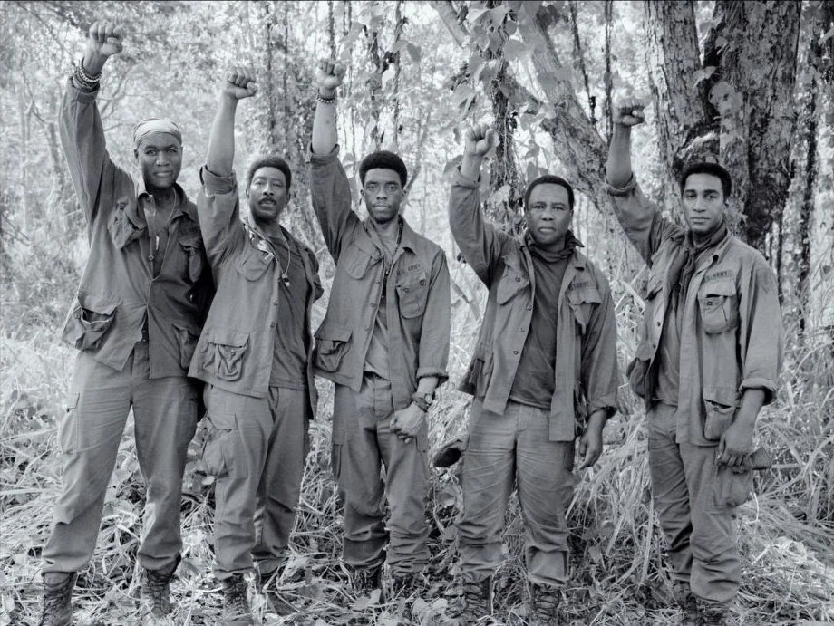 Da 5 Bloods (2020) movie still: fullbody — Five men in military uniform stand in a row within a dense jungle clearing. They hold their right arms…