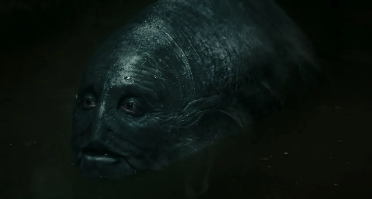 Pinocchio (2022) movie still: closeup — A dark, murky body of water contains a large, textured amphibian-like creature. The creature's head breaks…