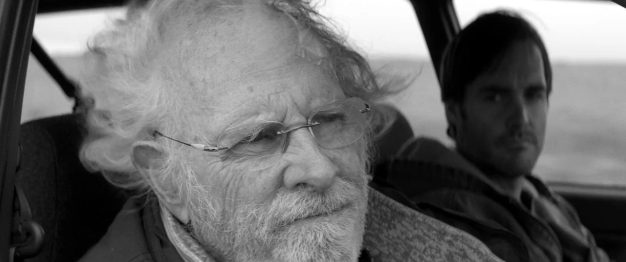 Nebraska (2013) movie still: medium — Two men are sitting in the front seats of a car during the day. The older man in the foreground is looking…