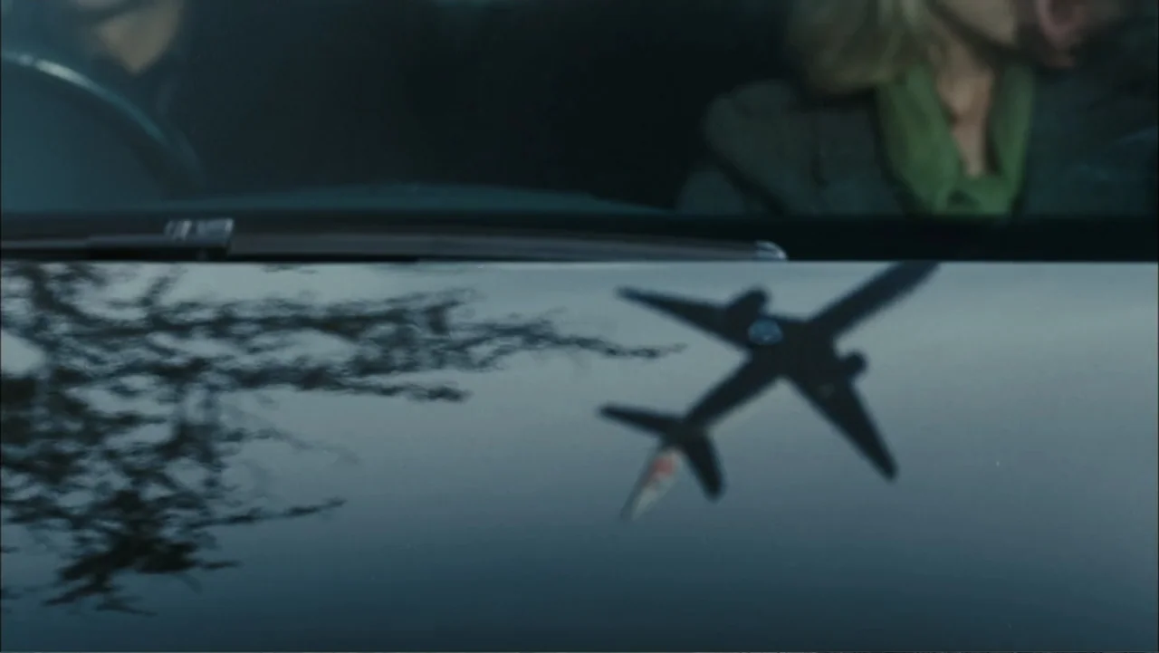 Before I Go to Sleep (2014) movie still: medium — A dark vehicle hood reflects the image of a flying passenger jet overhead. A driver and passenger…
