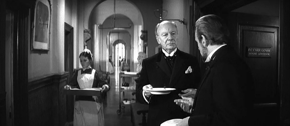 The Elephant Man (1980) movie still: medium — A long Victorian-era hospital hallway with vaulted ceilings and a sign on the door. Two men in formal…