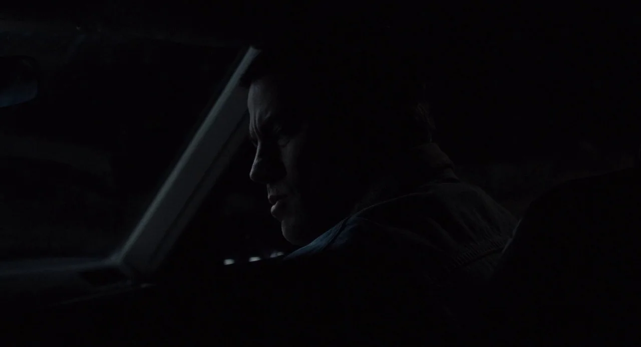 Foxcatcher (2014) movie still: medium — The interior of a dark car at night with minimal ambient light. A man sits in the driver seat looking out…