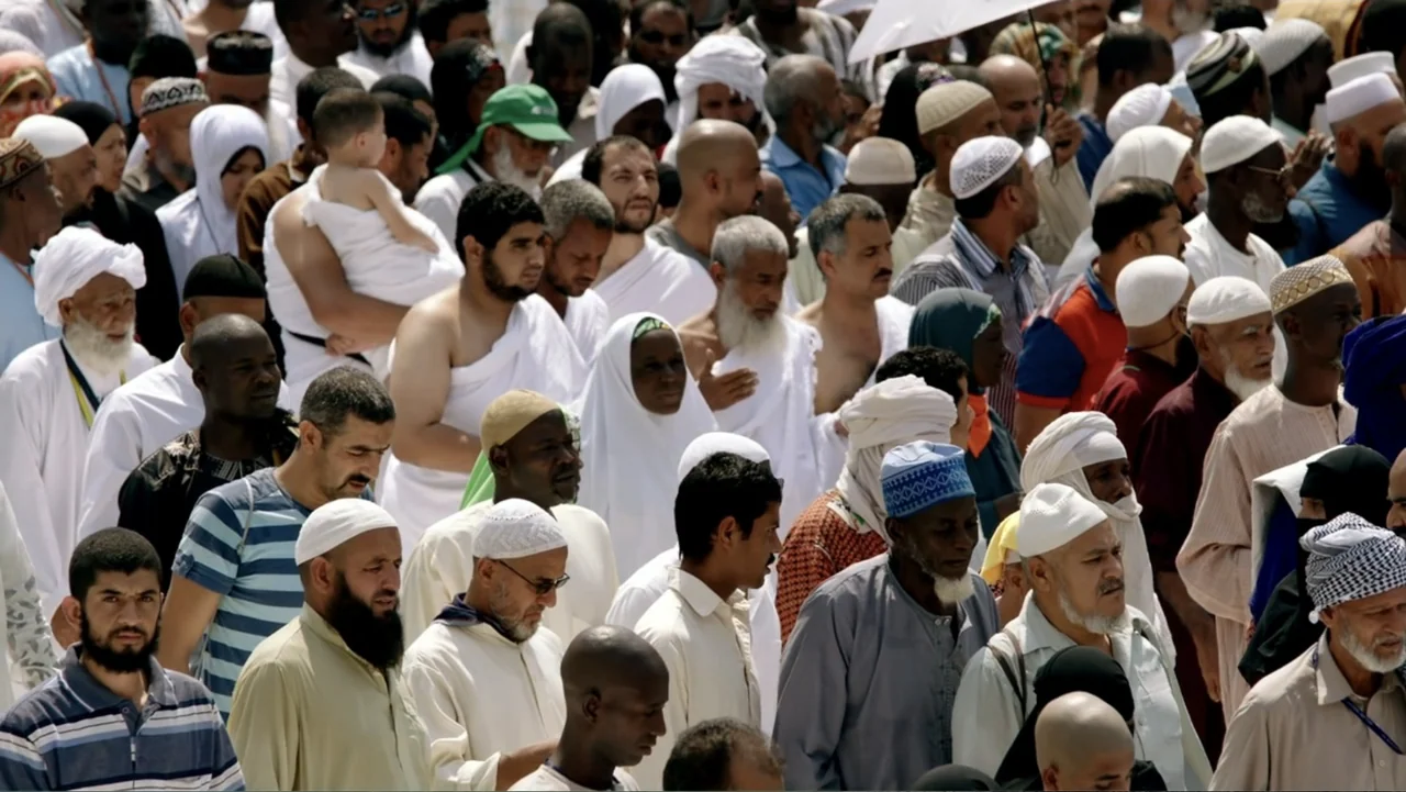 One Day in the Haram (2017) movie still: wide — A dense crowd of men in white religious garments standing in bright daylight. The scene shows a…