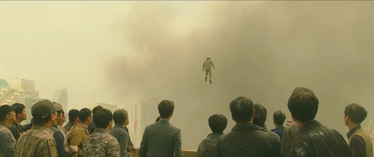Psychokinesis (2018) movie still: wide — A dense group of people stands in a hazy, yellowish urban environment looking upward. A lone figure is…