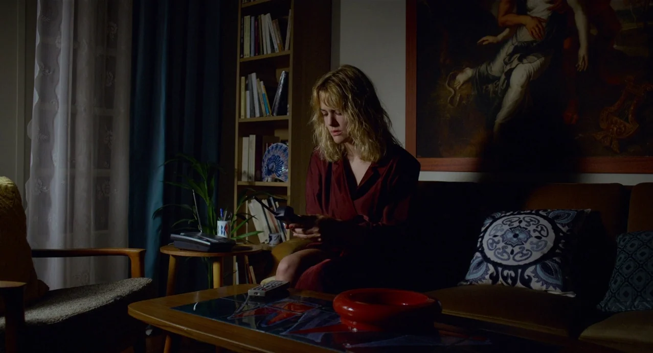Julieta (2016) movie still: medium — A dimly lit room with a bookshelf, a painting on the wall, and a sofa. A woman in a red robe sits on the sofa…
