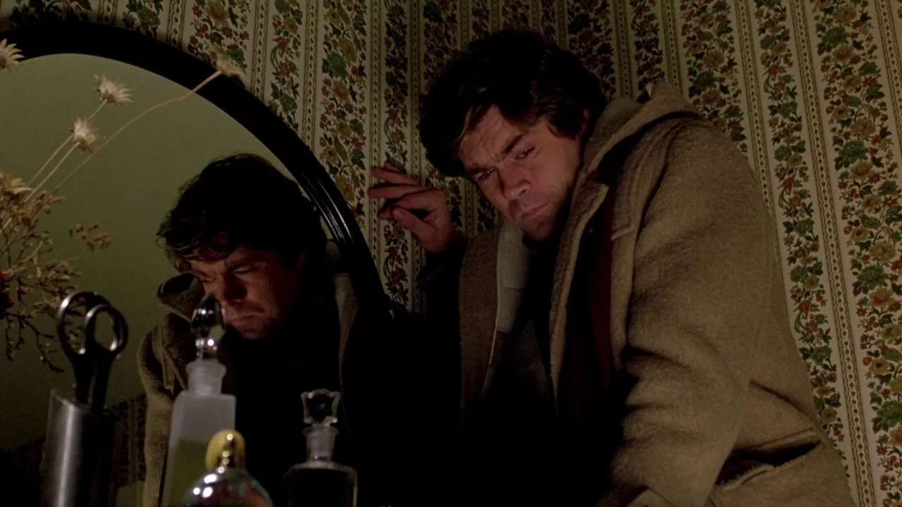 The Brood (1979) movie still: medium — A bathroom corner features dated floral wallpaper and a circular mirror with vanity items. A man in a wool…