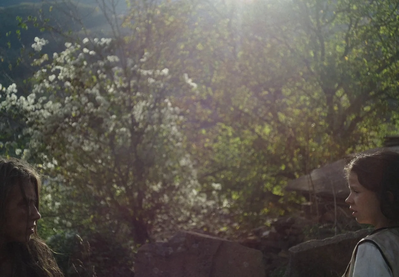You Won’t Be Alone (2022) movie still: medium — Two people stand in profile facing each other against a dense backdrop of trees. Sunlight glares…