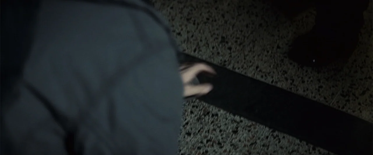 Hereafter (2010) movie still: closeup — A dark indoor space features a speckled aggregate floor and a blurred dark garment. A hand is visible…