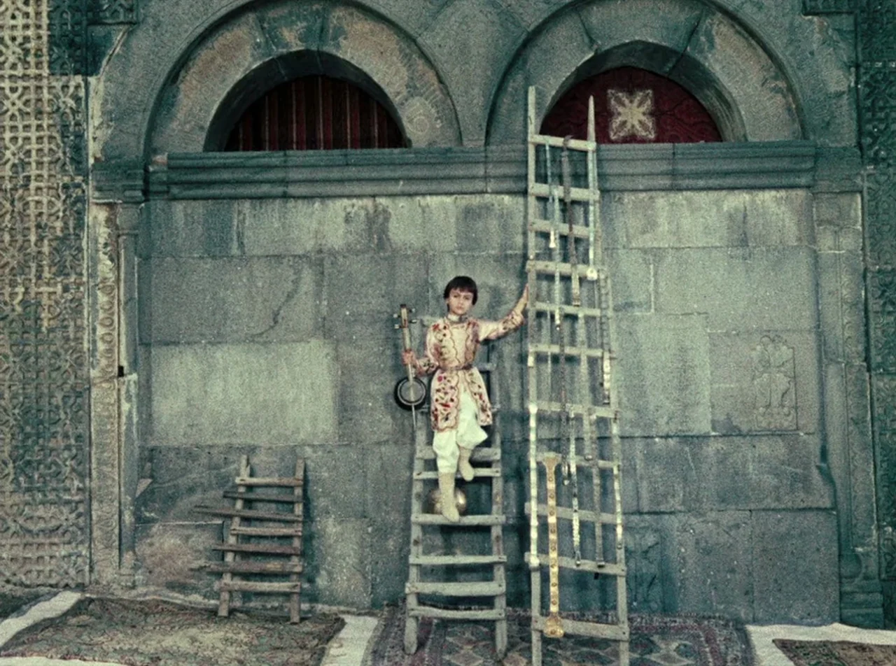 The Colour of Pomegranates (1969) movie still: fullbody — A child stands on a wooden ladder against a weathered gray stone wall. The wall features…