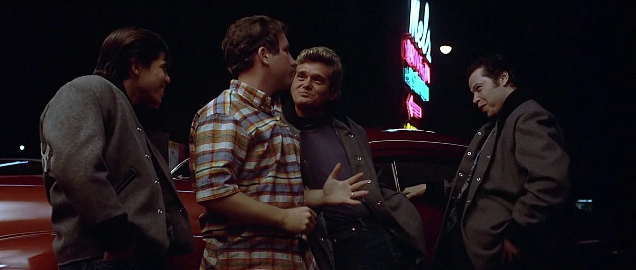 American Graffiti (1973) movie still: medium — Four men gather in a dark paved lot beneath a brightly illuminated vertical neon sign. They stand…