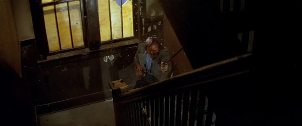 The Getaway (1972) movie still: medium — A dilapidated stairwell with dark wooden railings and debris-strewn floors. A middle-aged man in a grey…