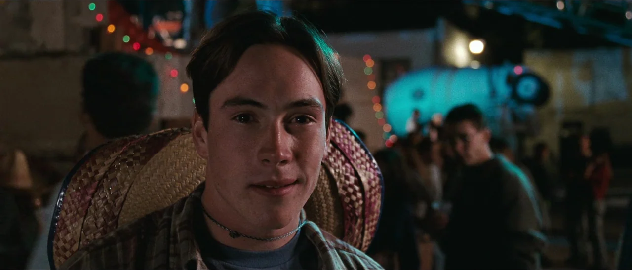 Election (1999) movie still: closeup — A nighttime outdoor party setting with blurred crowd members in the background. A young man sits in the…