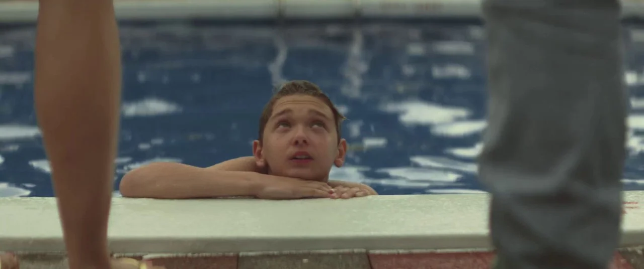 The Strange Ones (2017) movie still: medium — A tiled swimming pool deck with clear blue water and concrete coping. A teenage boy rests his chin on…