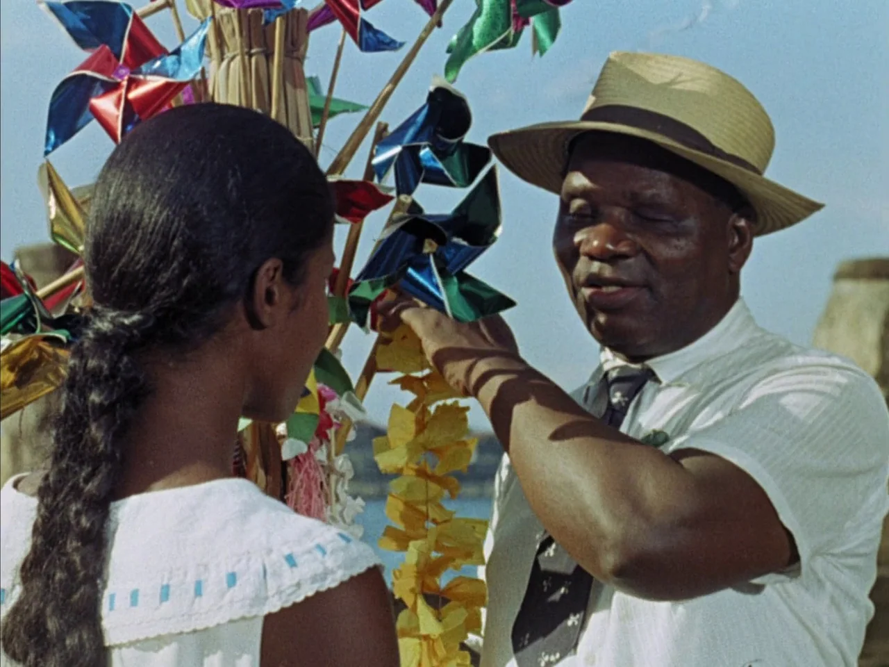 Black Orpheus (1959) movie still: medium — A sunny outdoor location features a man interacting with a cluster of bright pinwheels. An individual…