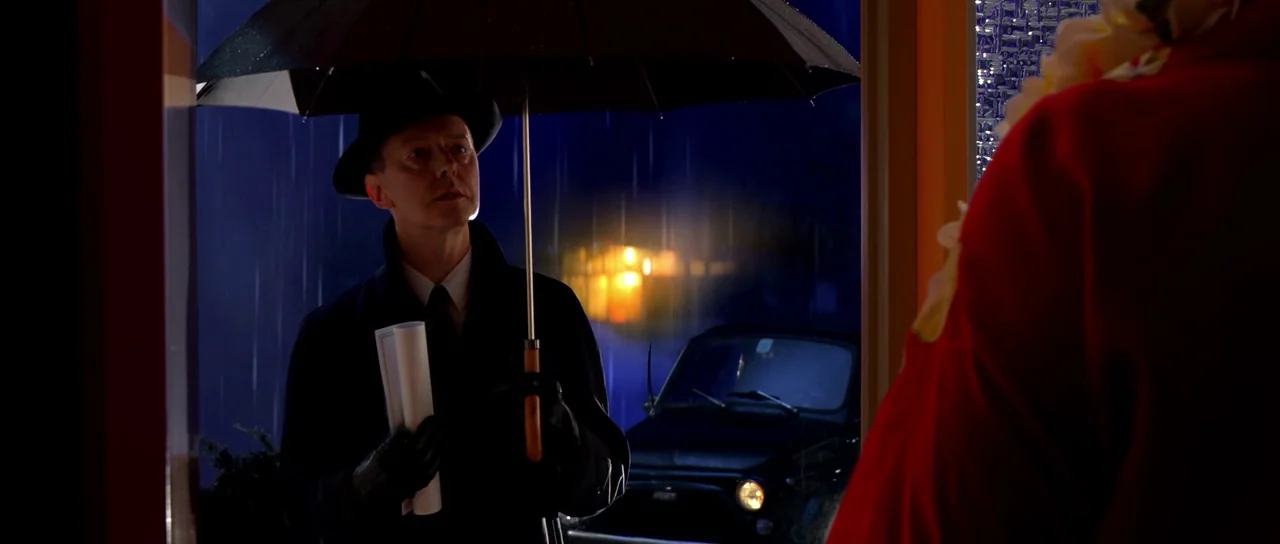 Speed Racer (2008) movie still: medium — A man in a fedora stands under an umbrella in the rain at night. A car is parked in the dark background lit…