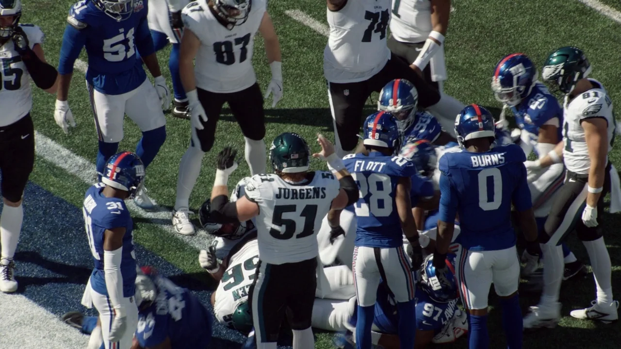 SAQUON (2025) movie still: wide — An American football field during a game with multiple players in team uniforms scattered. Several athletes in…