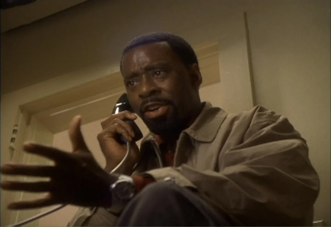 Ambushed (1998) movie still: medium — An interior space with pale walls and a white door frame. A man in a tan jacket is on a phone call while…