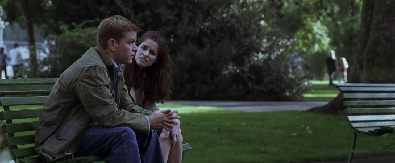 Syriana (2005) movie still: medium — Two people sit on a green wooden park bench in a verdant public space. A man and a woman face each other while…