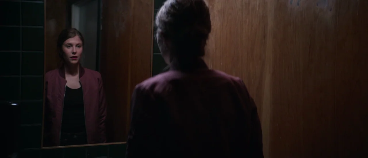Thelma (2017) movie still: medium — A dimly lit restroom with dark green tiled walls and wood paneling. A young woman stands looking at her own…