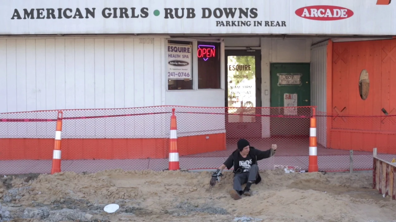 Buzzard (2014) movie still: wide — A commercial building front features signage for a health spa and arcade. A young man sits on a pile of dirt in…