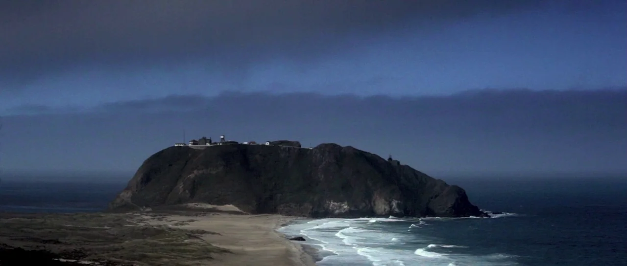 I Melt With You (2011) movie still: establishing — A rugged dark coastal cliff looms over a sandy beach under a cloudy sky. The ocean tide pulls…