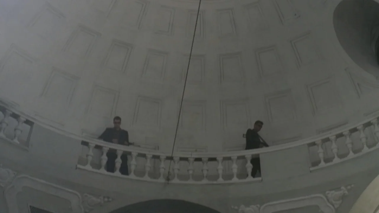 The Shepherd (2023) movie still: wide — A stone balcony inside a high domed rotunda features carved balustrades and paneled walls. Two men dressed…