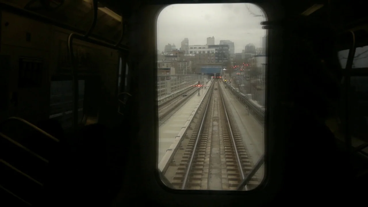 Counting (2015) movie still: wide — A view from the back of a moving subway car looking out over elevated tracks. Gray city buildings and overcast…