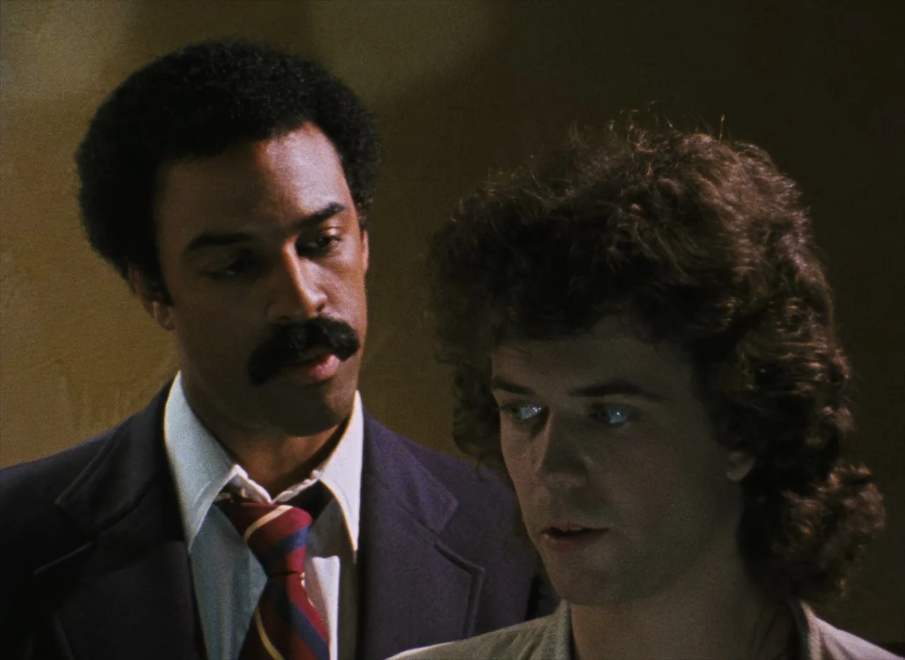 Basket Case (1982) movie still: medium — An interior setting features two men positioned closely against a muted dark background. The man on the…