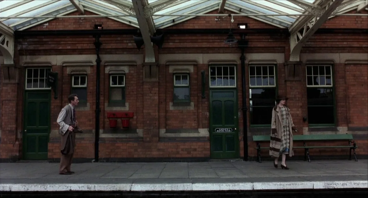 The Hours (2002) movie still: wide — An outdoor brick railway platform features green wooden doors and iron roofing support beams. A man and a woman…