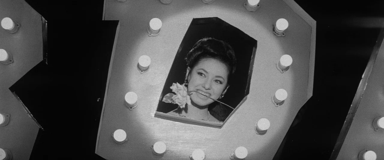 Carmen From Kawachi (1966) movie still: closeup — A woman appears through a large cutout lettered frame illuminated by numerous round bulbs. She is…