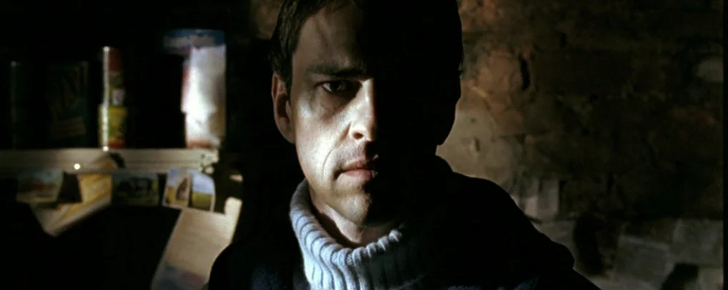 Calvaire (2004) movie still: closeup — A dimly lit stone-walled interior with shelves holding canned goods in the background. A man stands in the…
