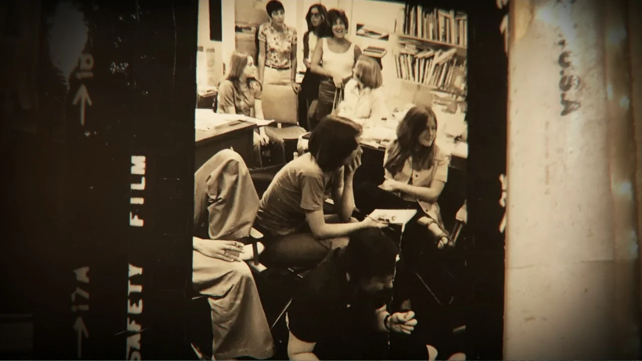 Dear Ms.: A Revolution in Print (2025) movie still: medium — A cluttered 1970s office contains several women sitting among bookshelves and desks.…