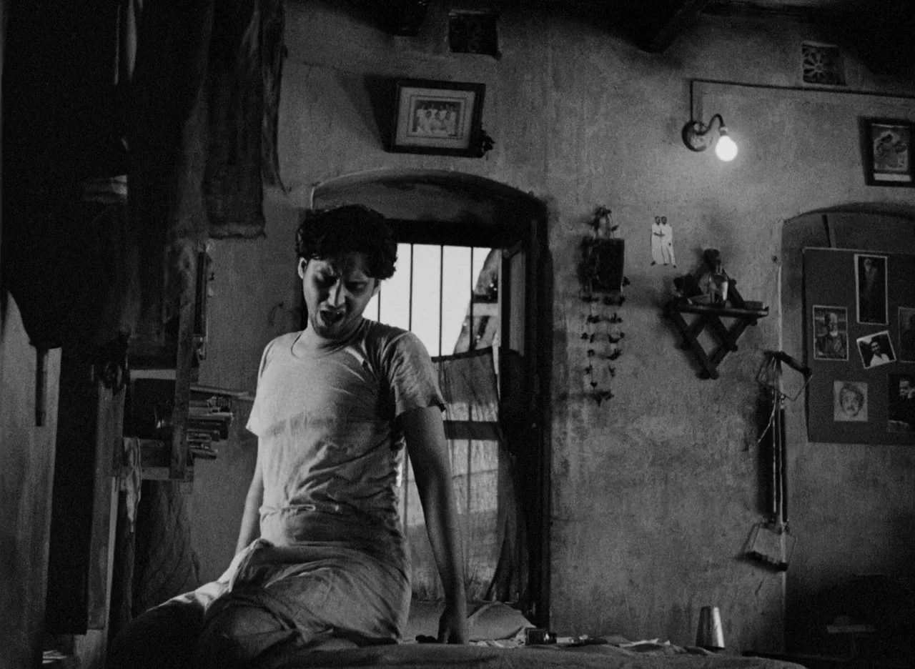 Apur Sansar (1959) movie still: medium — A dimly lit room features textured walls adorned with framed pictures and a small lamp. A young man yawns…