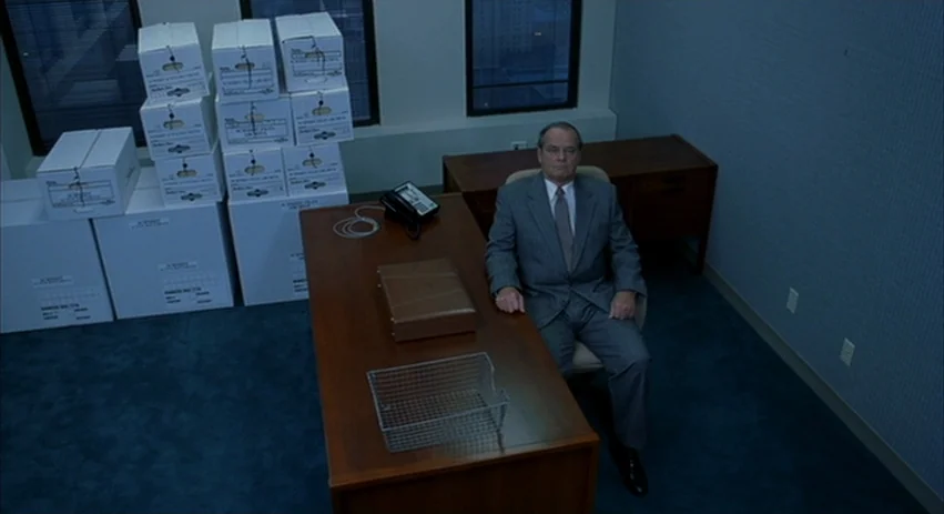 About Schmidt movie still: wide — A sparsely furnished room features blue carpeting and rows of stacked white archive boxes. A middle-aged man in a…