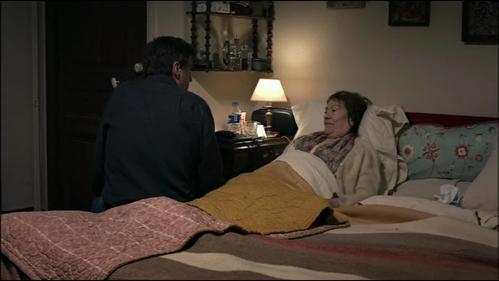 Caché (Hidden) (2005) movie still: medium — A dimly lit bedroom features an elderly woman reclining in a bed with blankets. A man sits beside her at…