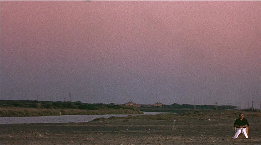 Juliet of the Spirits (1965) movie still: wide — A man sits alone on a folding chair in an expansive, flat coastal landscape. A calm river or inlet…