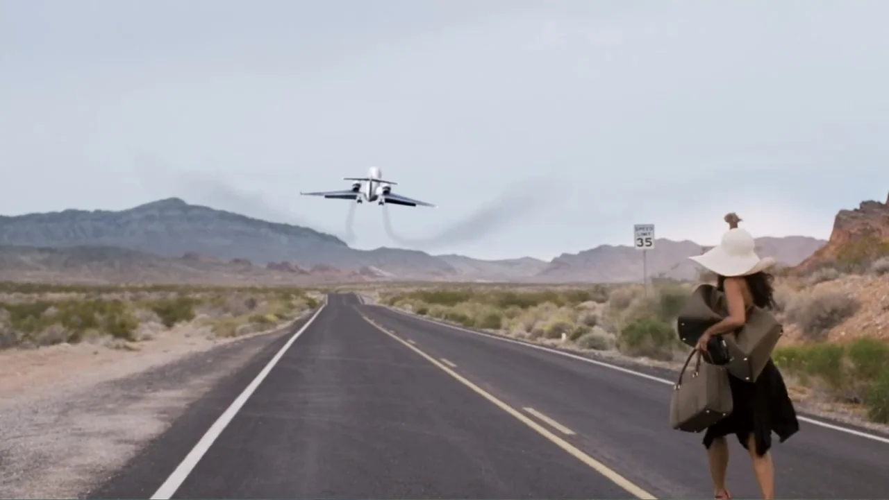 Redline (2007) movie still: wide — A flat desert highway extends into the distance toward low mountains under overcast sky. A woman in a black dress…
