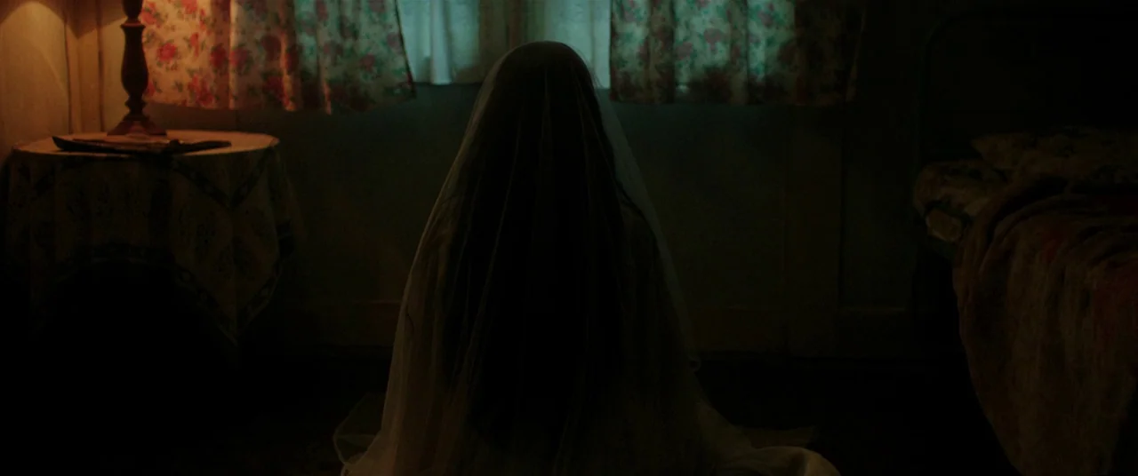 Satan’s Slaves (2017) movie still: medium — A dimly lit room features a person covered in a white veil sitting centrally. Floral curtains flank the…