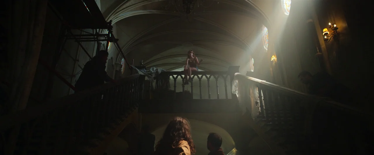The Pope’s Exorcist (2023) movie still: wide — A dark vaulted stairwell inside an older building features heavy wooden railings and arches. A woman…