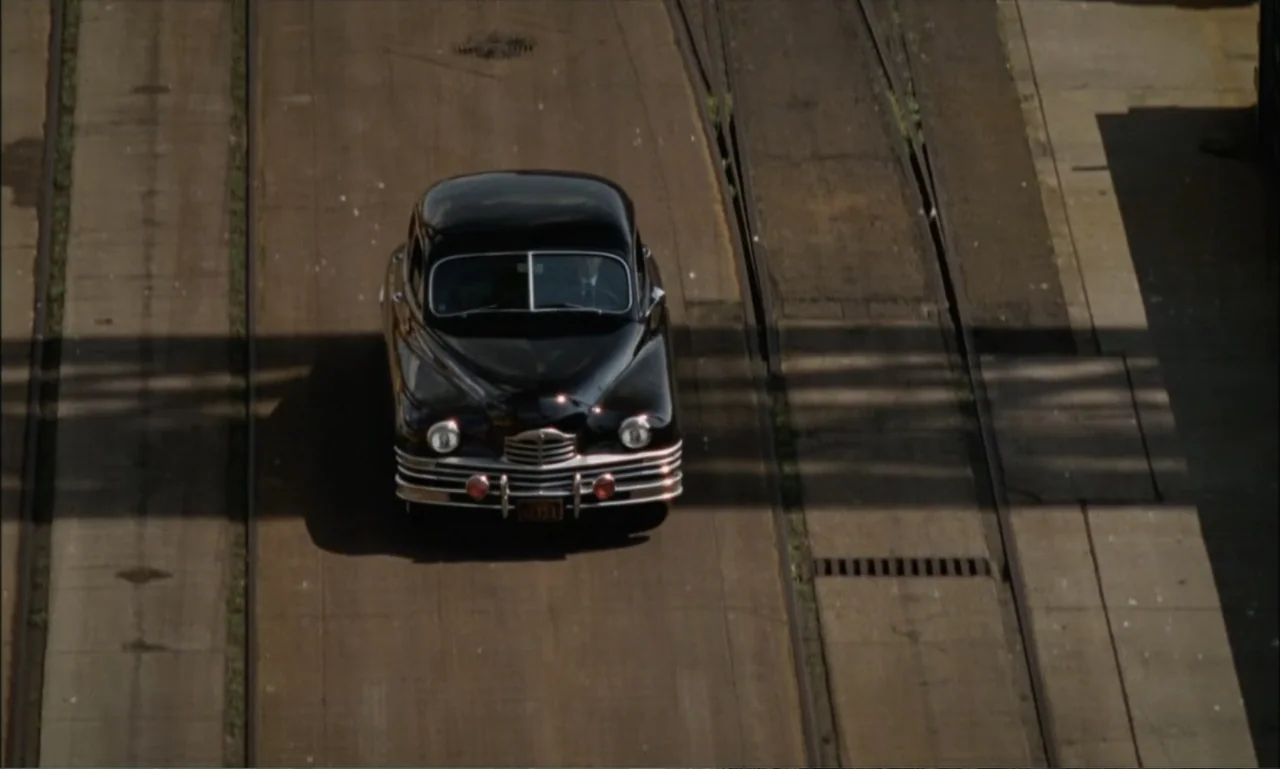 The Brutalist (2024) movie still: wide — A dark vintage car drives on a paved area featuring embedded railroad tracks. The scene is captured from an…