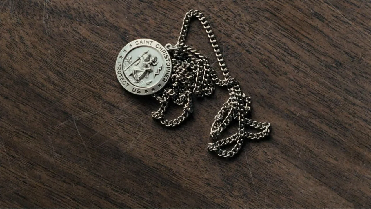 Taking Chances (2011) movie still: closeup — A Saint Christopher medal with a silver chain rests on a dark wood surface. The surface has faint…