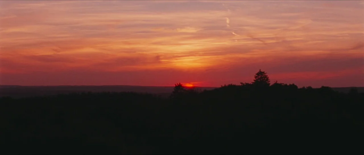 Inexorable (2021) movie still: establishing — A vast landscape shows a sunset against a distant, silhouetted treeline and flat horizon. The bright…