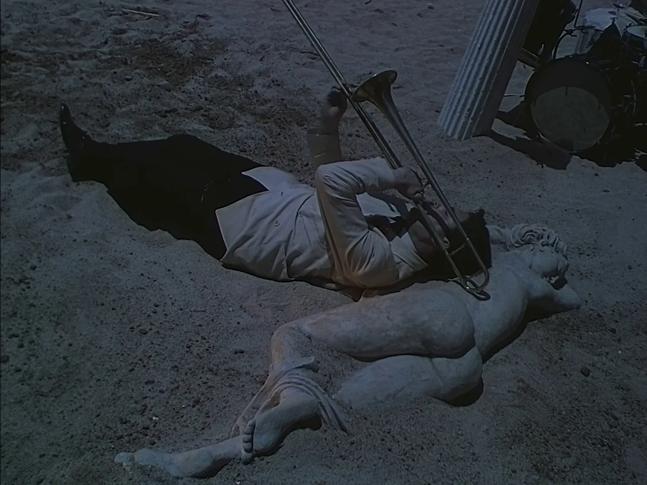 Pandora and the Flying Dutchman (1951) movie still: fullbody — A man lies on a sandy beach at night next to a reclining stone statue. He plays a…
