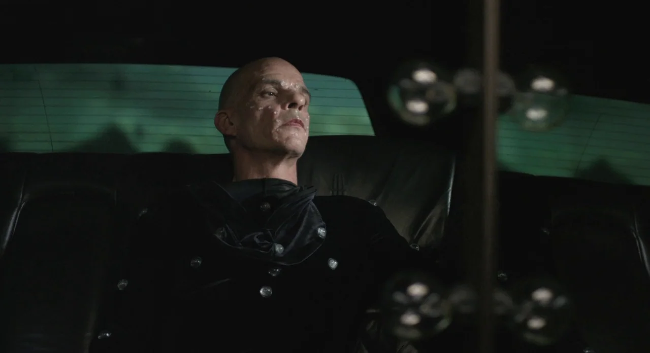 Holy Motors (2012) movie still: medium — A man sits in the dark back seat of a car at night. The view through the rear window shows a blurry green…