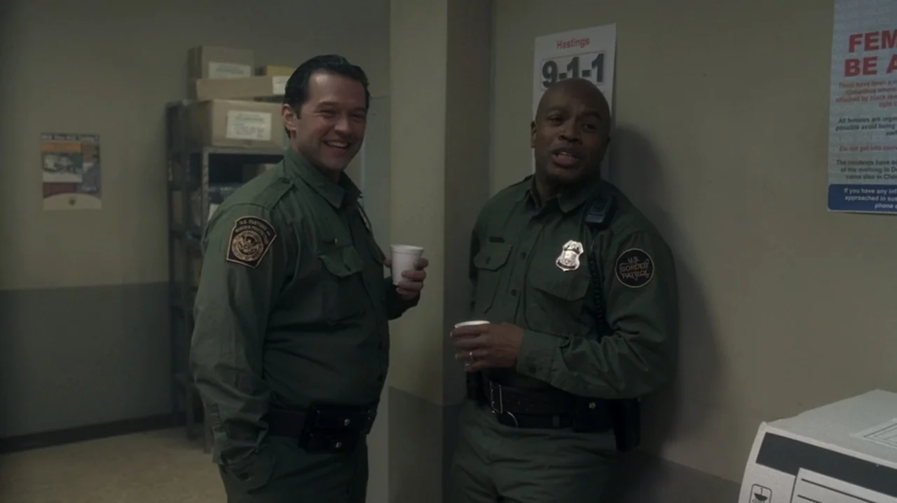 The Shepherd (2023) movie still: medium — A utilitarian office space has beige walls decorated with official government warning posters. Two male…
