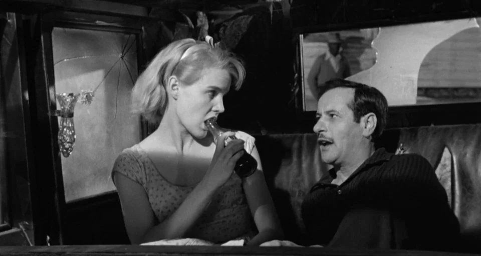 Baby Doll (1956) movie still: medium — A cramped car interior features cracked windows and worn leather upholstery. A blonde woman drinks from a…